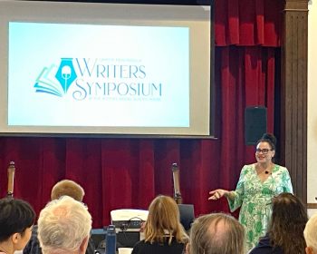 UPPAA A person wearing glasses and a green dress is speaking on stage at the Upper Peninsula Writers' Symposium. The event logo is displayed on a screen behind them. The audience is seated, facing the stage.